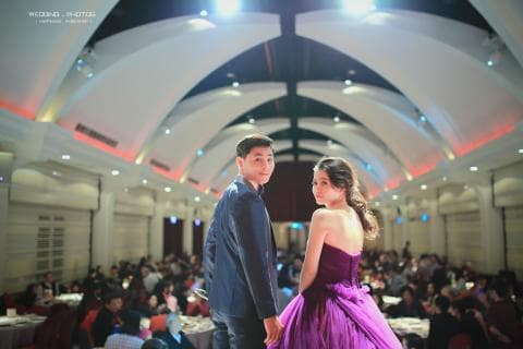 這張照片是一對新人在婚禮現場，新郎穿著深藍色西裝，新娘穿著紫色的婚紗，他們互相依偎，臉上洋溢著幸福的笑容，背景是一片歡樂的婚禮場景。