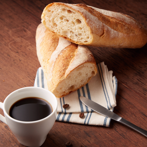 這張「麵包、咖啡和刀子」的照片是，兩個法式長棍麵包放在藍白條紋的餐巾紙上，旁邊放了一杯咖啡和一把刀子。