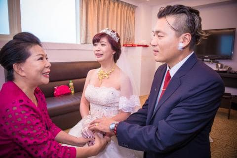 這張照片是一個婚禮的場景，照片中新郎正為新娘戴上戒指，新娘的親朋好友在一旁觀禮，場面溫馨。