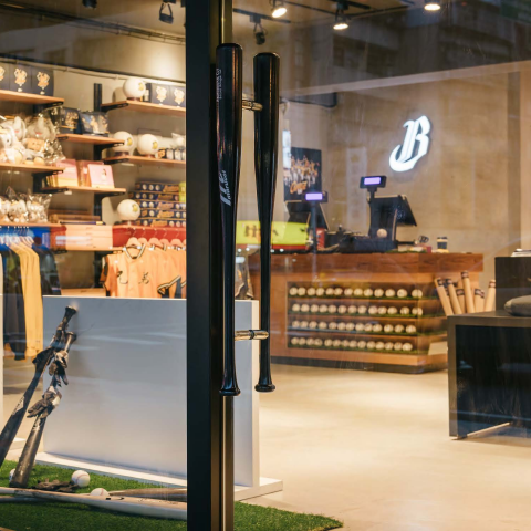 這張「棒球用品店」的照片是一家裝潢時尚的商店，店內擺放著各種棒球用品，包括球棒、球和帽子。牆上掛著一排棒球裝備，店裡還有玻璃櫃展示各種棒球相關的商品。門上裝了兩個黑色棒球棍形門把，很有特色。