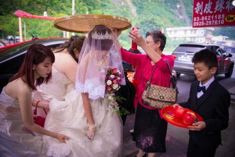 這張照片是一個婚禮，新娘穿著白色的婚紗，頭戴著頭紗，新郎站在新娘旁邊，他穿著黑色的西裝，新娘的母親穿著紅色的洋裝，手拿著一個禮物，新娘的妹妹穿著粉紅色的洋裝，手拿著一個花束，新娘的父親站在後方，手拿著一個相機。