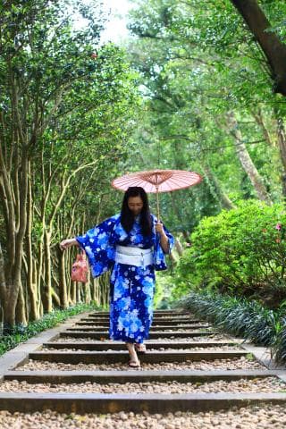 這張照片是一位身穿藍色花紋和服的女性，她正走在石階上，手裡拿著一把粉紅色的雨傘，她的表情看起來很開心。