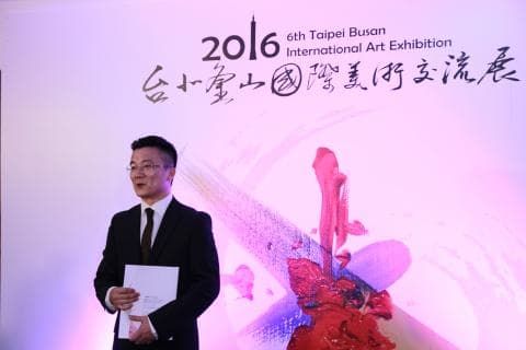 這張「2016 臺北釜山國際藝術展」的照片是一位男士站在粉紅色背景前，背景上有一幅抽象畫。