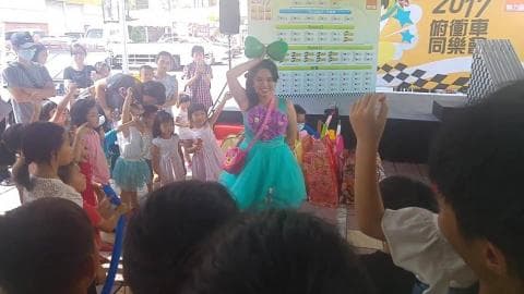 這張照片是一個舞臺表演，一位女演員穿著粉藍色的洋裝，頭上戴著綠色的頭飾，正在臺上表演，臺下有很多小朋友在觀看。