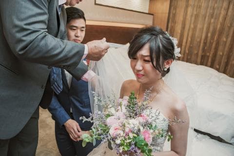 這張「婚禮」的照片是一個新郎、新娘和一位長輩。新娘穿著白紗，頭戴頭紗，手裡拿著花束，看起來很幸福。長輩則替她蓋著頭紗，表情溫馨。新郎站在新娘旁邊，露出笑容。