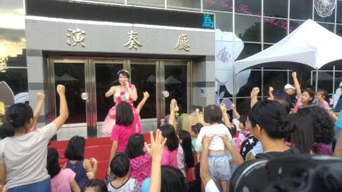 這張「兒童表演」的照片是，一位女歌手在臺上唱歌，臺下許多小朋友開心地揮手跟著唱歌。