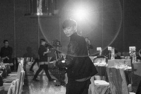 這張照片是一個穿著外套的年輕人手持相機站在一群人中間，旁邊有桌子和椅子。