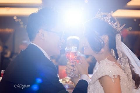 這張婚禮的照片是一對新人舉杯相碰，新娘穿著白紗，頭戴皇冠，新郎穿著西裝。