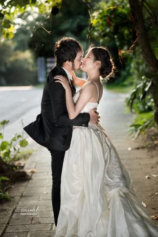 這張婚禮照片是一對新人互相擁吻，新郎穿著黑色西裝，新娘穿著白色婚紗，他們在戶外綠樹環繞的地方，看起來非常甜蜜幸福。