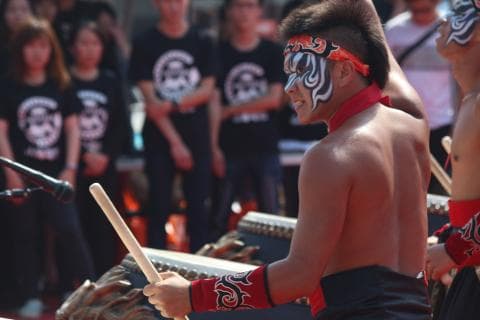 這張「表演者」的照片是一個畫著臉譜的男子正在敲打一個大鼓，旁邊圍觀著一群觀眾。