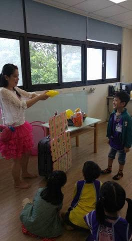 這張照片是一群小朋友在玩遊戲，一位年輕女性正在為他們主持。最顯眼的是中間的遊戲道具，是一個由許多格子的牌組成的立體遊戲，每個格子都印著不同的圖案，旁邊放著一個行李箱和一些禮物。