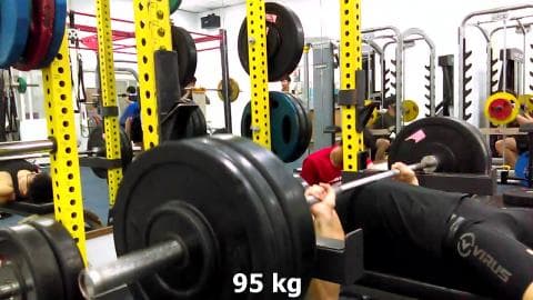 這張照片是一名男子在健身房做臥推運動。他正在舉起 95 公斤的槓鈴，臉上展現出專注的神情。