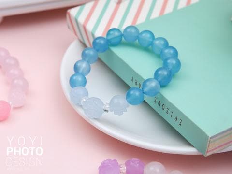 這張「藍色珠子手環」的照片是放在一本綠色封面的筆記本上，手環上有兩個小飾品，其中一個是象，另一個是花朵。