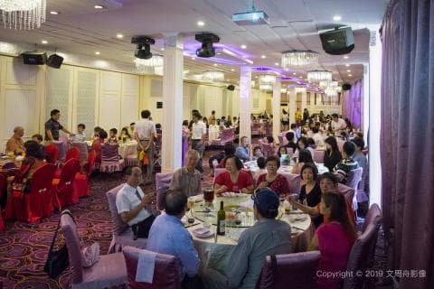 這張照片是一場婚宴，賓客們正圍坐在餐桌旁享用美食，桌上擺滿了豐盛的菜餚，氣氛熱鬧而溫馨。