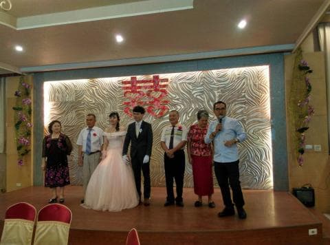 這張照片是一對新人舉辦婚禮的現場，新娘穿著白色婚紗，新郎穿著黑色西裝，他們正準備交換戒指。新郎的父母和親友們站在一旁觀禮，場面溫馨而喜悅。