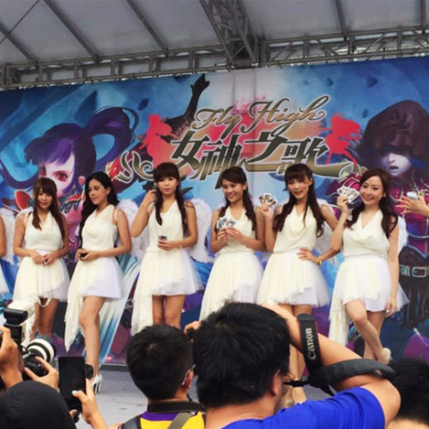 這張「Fly High 女神」的照片是六個穿著白色洋裝的女生，站在舞臺上，她們手拿卡片，背景是遊戲的宣傳海報。