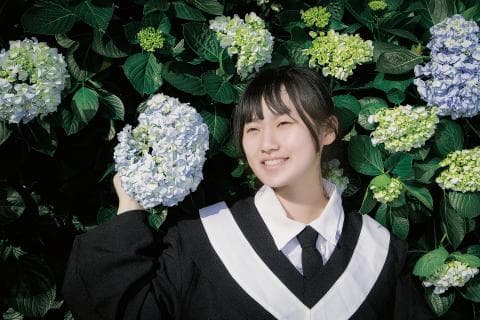 這張「一位年輕女子站在花叢中」的照片是一個年輕女子站在花叢中的照片。她穿著一件黑色的制服，並且正在看著鏡頭。 她的表情是微笑的，並且她看起來很開心。 她手中拿著一朵白色的花，並且周圍有很多綠色的植物。