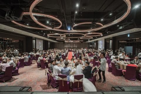 這張婚禮現場的照片是一場正在舉行的婚禮宴會，佈置華麗，許多賓客圍坐在一起，場面熱鬧。舞臺上有一對新人正在舉行儀式，他們身穿婚紗禮服，背景是華麗的燈光和裝飾。