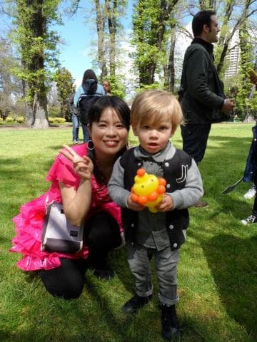 這張「一家三口在公園裡玩耍」的照片是一名穿著粉紅色衣服的女子抱著一個小男孩，小男孩手裡拿著一個黃色的玩具，女子旁邊站著一位男子。