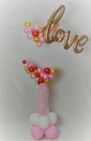 這張氣球裝飾的照片是一個愛心形狀的氣球拱門，上面有金色 love 字樣氣球和粉色、紅色、金色的小氣球，底部有白色和粉色的氣球，以及粉色薄紗。