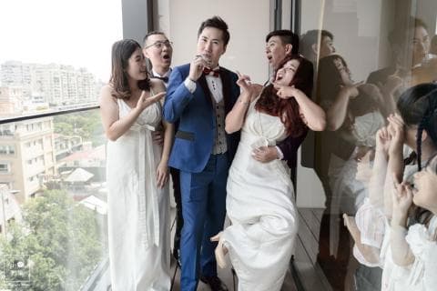 這張照片是一對新人在結婚當天，與親朋好友們一起歡樂的畫面。他們站在窗邊，看著外面的風景，新郎穿著藍色的西裝，新娘穿著白色的婚紗，他們的手裡都拿著酒杯，臉上都洋溢著幸福的笑容。他們看起來很開心，也充滿了愛。