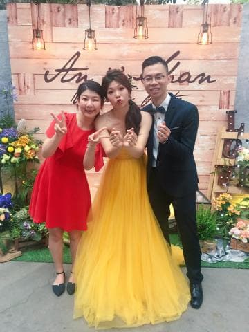 這張婚禮的照片是兩個女人和一個男人在木板牆前合影。  一個女人穿著紅色禮服，另一個女人穿著黃色婚紗，男人則穿著西裝。