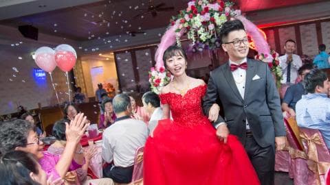 這張照片是一對新人舉行婚禮，新娘穿著紅色禮服，新郎穿著黑色西裝，兩人牽手走進禮堂，背景是花拱門和氣球，畫面充滿幸福和喜悅。