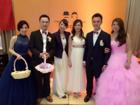 這張照片是一對新人在婚禮上拍的照片，照片中的新人穿著西裝禮服，新娘穿著白色的婚紗，他們站在一起並手持花籃，周圍有親朋好友陪伴，場面十分喜慶。