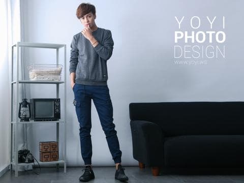 這張「時尚」的照片是一個年輕男子穿著灰色毛衣和深藍色長褲，站在白色背景的房間裡，他左手放在下巴上，右手指著前方。他身後有一個黑色的沙發，牆上有一個白色字體的品牌標誌。