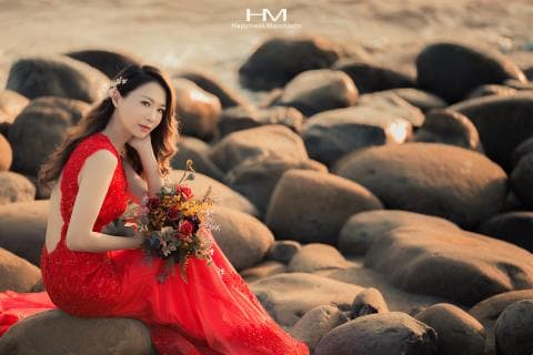 這張「婚紗照」的照片是一位穿著紅色婚紗的女子，坐在海邊的岩石上，手裡拿著一束鮮花，背景是許多大小不一的石頭。