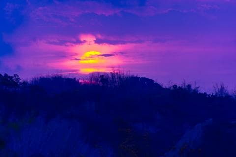 這張「夕陽」的照片是黃色的太陽從紫色的雲層中落下，遠處的山丘呈現藍黑色的剪影。