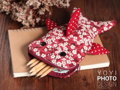 這張「鯊魚造型鉛筆盒」的照片是，一個用紅色布料縫製而成的鯊魚形狀鉛筆盒，裡面裝滿了鉛筆，旁邊還有一本筆記本。