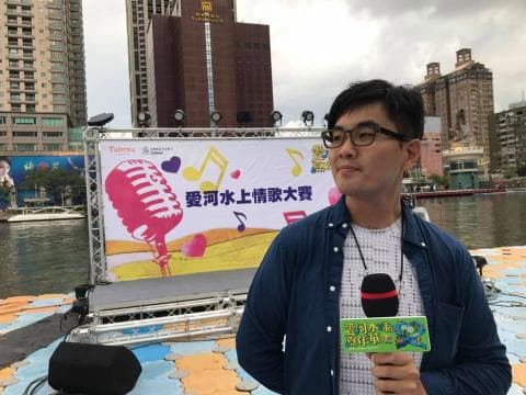 這張「愛河水上情人節」的照片是，一位男士站在舞臺前，拿著麥克風，準備唱歌。舞臺上有一個大型的看板，上面有愛河水上情人節的標誌，以及一個巨大的麥克風。