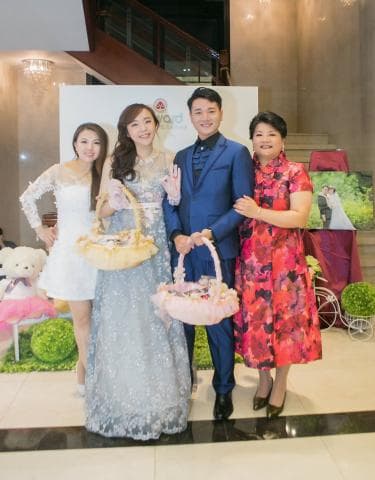 這張照片是三位女性和一位男性站在一起，其中一位女性穿著白色蕾絲連衣裙，另一位女性穿著藍色亮片禮服，第三位女性穿著紅色花卉圖案禮服，男性穿著藍色西裝。他們都帶著笑容，看起來很開心。