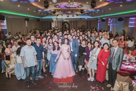 這張「婚禮」的照片是一對新人和他們的親朋好友在婚宴上合影。 新娘穿著粉紅色的婚紗，新郎穿著西裝，他們站在中間， 周圍是許多賓客。 他們都笑得很開心， 畫面充滿著喜悅和祝福。