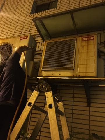 這張照片是一個工人正在修理一台室外冷氣機，他站在梯子上，並使用工具來檢查冷氣機。