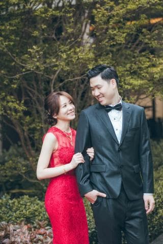 這張照片是一對新人在綠樹叢中拍攝婚紗照，照片中主要呈現了新郎穿著黑色西裝，新娘穿著紅色禮服，他們互相依偎，眼神充滿愛意。