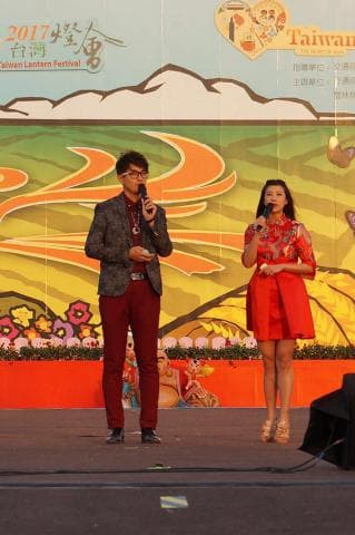 這張照片是臺灣燈會活動的舞臺，兩位主持人正站在舞臺上。舞臺背景是黃色和橘色的，代表著喜慶的氛圍。舞臺上還擺放了一些裝飾，增添了活動的色彩。