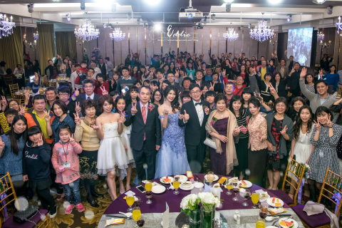 這張照片是新郎新娘和他們的親朋好友在婚宴上合照。新郎新娘站在中間，他們都穿著正式的禮服。新郎穿著黑色的西裝，新娘穿著藍色的禮服。他們的身後是他們的家人和朋友，他們都面帶笑容，看起來非常開心。