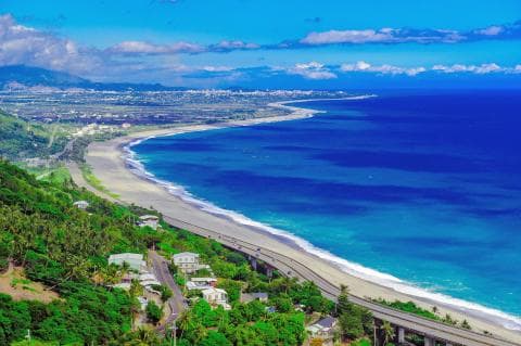 這張「海岸公路」的照片是 一條蜿蜒的公路沿著海岸線延伸，公路旁是沙灘和碧藍的海水，遠處是綿延的山脈。