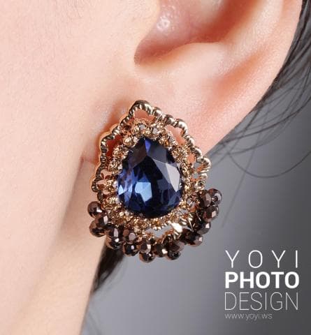 這張照片是一個戴著耳環的女性耳朵的特寫。耳環的中心是一顆藍色的水滴形寶石，周圍環繞著金色的花朵和水晶。