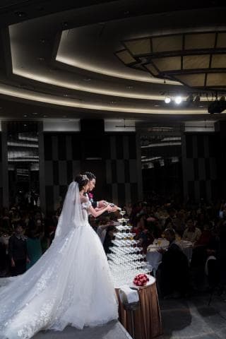 這張婚禮的照片是一對新人正在用香檳酒杯塔進行婚禮儀式。 新娘穿著白色婚紗，新郎穿著西裝，他們手持酒瓶，準備將香檳倒入酒杯塔。  背景是婚禮宴會廳，有很多賓客圍觀。