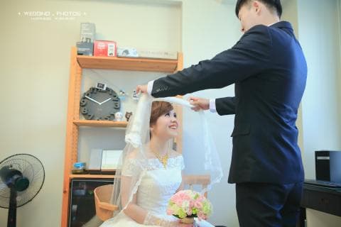 這張照片是婚禮中新郎為新娘蓋頭紗的溫馨瞬間。新娘穿著美麗的婚紗，新郎則身穿西裝，神情專注地為新娘蓋頭紗，動作輕柔，充滿愛意。頭紗飄逸，營造出浪漫的氛圍。