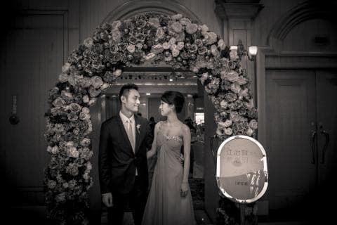 這張照片是一對新人站在花門下，即將步入婚姻的殿堂。新郎身穿西裝，新娘則穿著婚紗，他們深情地凝視著彼此，臉上洋溢著幸福的笑容。