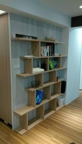 這張照片是一個現代化的開放式書架，它由許多個木質隔板組成，並在牆上建立了一個獨特的幾何形狀。 書架上有各種物品，例如書籍、裝飾品和日常生活用品。 整個設計簡潔時尚，與背景的淺色牆壁搭配得很好，為空間增添了一份現代感。 
