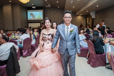 這張照片是一對新人在婚禮宴會上，新郎穿著灰色西裝，新娘穿著粉紅色禮服，他們手牽手，幸福地微笑著。