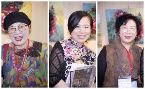 這張「三個人站在藝術畫作前」的照片是，左邊是一位戴著帽子、穿著花色衣服的女性，中間是一位穿著花色洋裝、拿著書的女性，右邊是一位穿著棕色外套的女性。