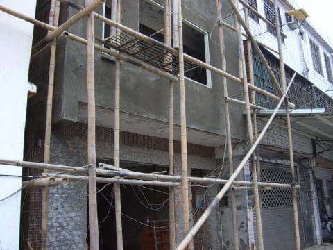這張照片是一棟正在修建的建築物，它被大量的竹製鷹架包圍著。鷹架從地面一直延伸到屋頂，上面掛著許多電線和管道。建築物的牆壁上還有一扇窗戶，窗戶上也有一層鷹架。看起來這棟建築物正在進行翻新工程。