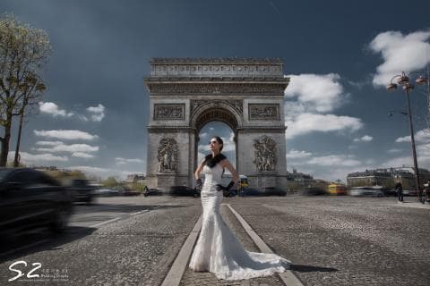 這張照片是一個美麗的新娘站在巴黎凱旋門前，她穿著優雅的婚紗，頭上戴著花環，手裡拿著一束花，臉上洋溢著幸福的笑容。