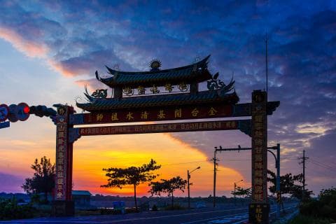 這張「中國傳統牌坊」的照片是日出時，有著美麗的橘紅色天空。牌坊上寫著「歡迎光臨」四個字，下方有兩條道路，路邊還有許多樹木。